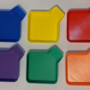 May include: Six brightly coloured, square trays with a distinctive angled corner design. The trays are in blue, yellow, red, purple, green, and orange. The trays are likely plastic and are arranged on a white surface.