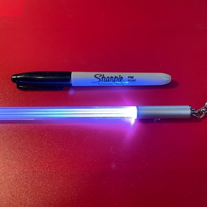 Peut inclure: Un porte-cl&eacute;s argent&eacute; avec un tube lumineux transparent, blanc et bleu. Le porte-cl&eacute;s est sur une surface rouge &agrave; c&ocirc;t&eacute; d'un marqueur permanent Sharpie noir et blanc &agrave; pointe fine.