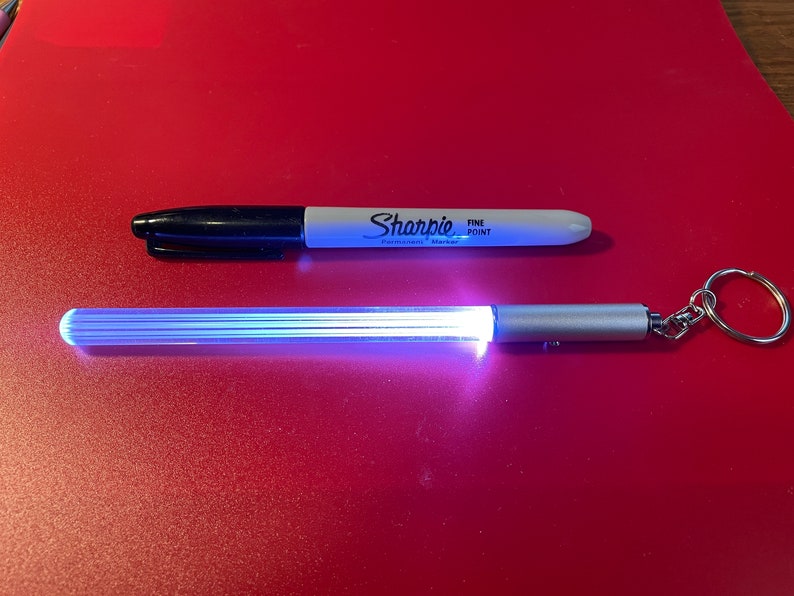 Peut inclure: Un porte-cl&eacute;s argent&eacute; avec un tube lumineux transparent, blanc et bleu. Le porte-cl&eacute;s est sur une surface rouge &agrave; c&ocirc;t&eacute; d'un marqueur permanent Sharpie noir et blanc &agrave; pointe fine.