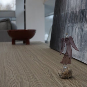 May include: A glass angel figurine with a purple iridescent finish stands on a small, gray rock. The angel is facing away from the camera, with its wings spread out behind it. The figurine is on a wooden table.