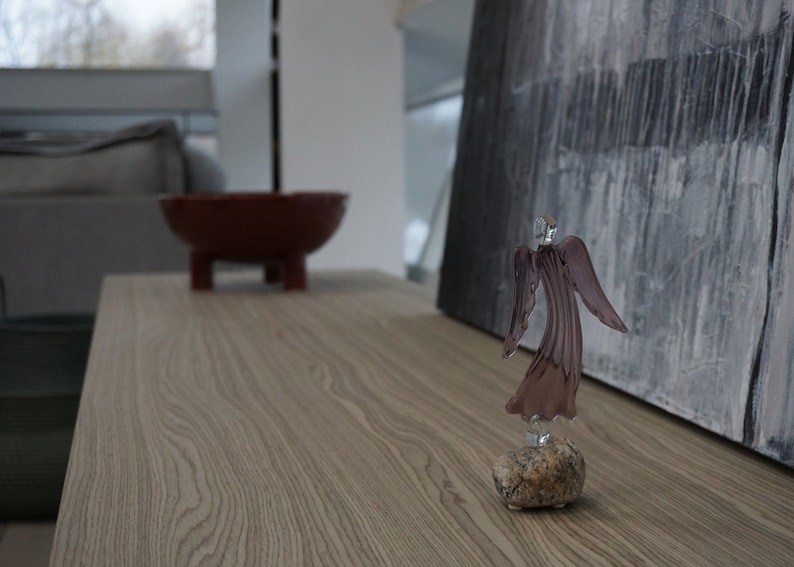 May include: A glass angel figurine with a purple iridescent finish stands on a small, gray rock. The angel is facing away from the camera, with its wings spread out behind it. The figurine is on a wooden table.