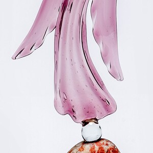 May include: A pink glass angel figurine standing on a brown and red stone base.