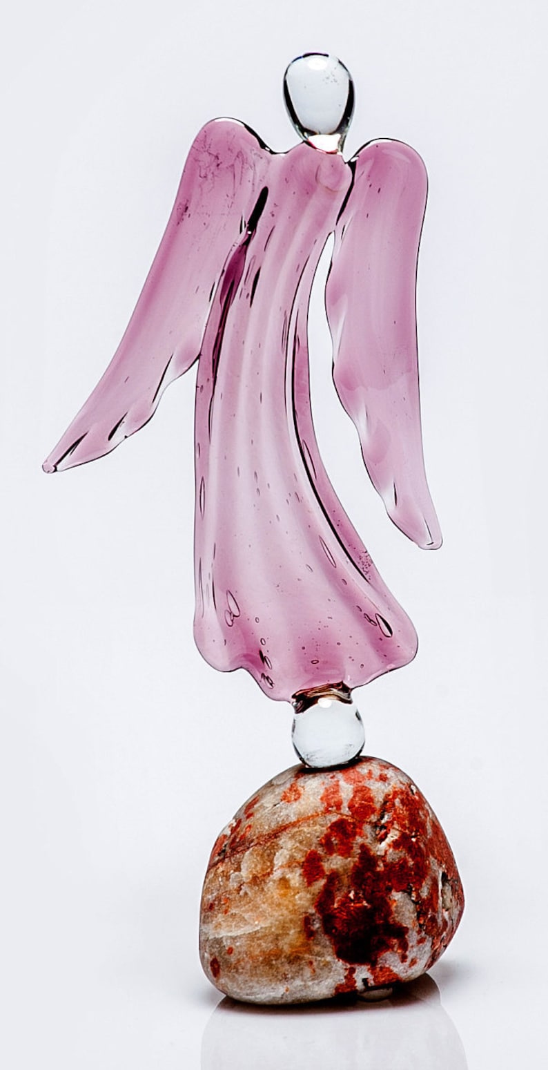 May include: A pink glass angel figurine standing on a brown and red stone base.