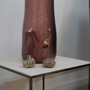 May include: Two gold glass bird sculptures on a white square table with a textured purple vase in the background.