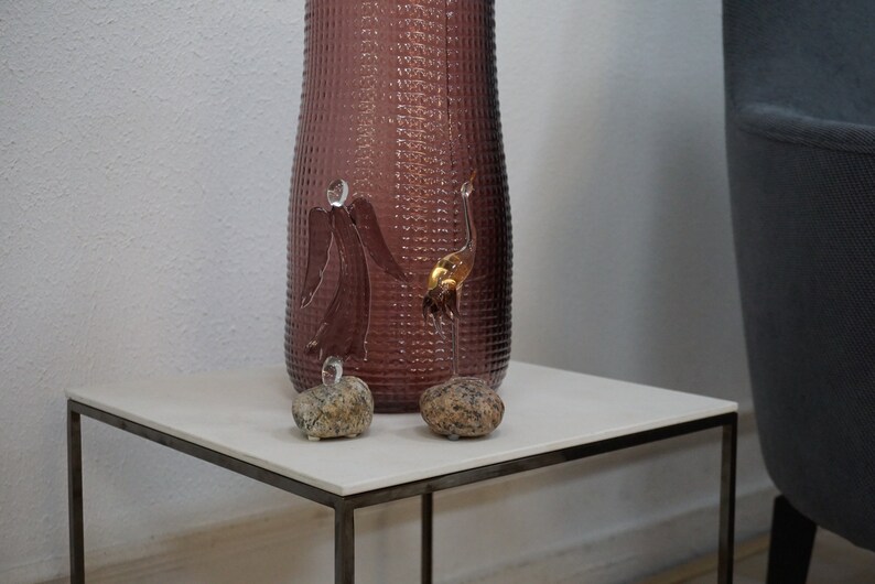 May include: Two gold glass bird sculptures on a white square table with a textured purple vase in the background.