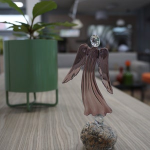 May include: A glass angel figurine with a pink iridescent finish stands on a small gray stone. The angel has outstretched wings and a delicate, flowing gown. The figurine is on a wooden table with a green potted plant in the background.