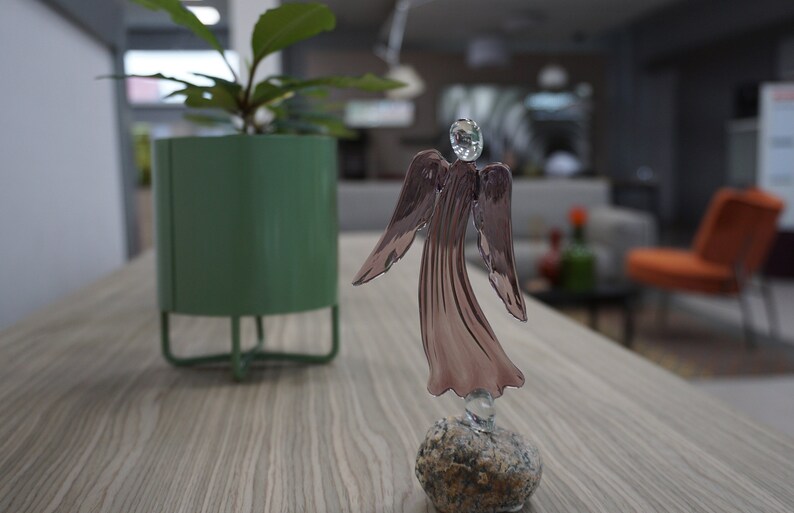 May include: A glass angel figurine with a pink iridescent finish stands on a small gray stone. The angel has outstretched wings and a delicate, flowing gown. The figurine is on a wooden table with a green potted plant in the background.