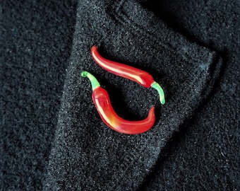 May include: Two red chili peppers with green stems are pinned to a black fabric surface.