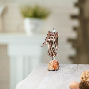 May include: A glass angel figurine with a rose gold finish standing on a small, red stone. The angel is standing on a wooden surface.