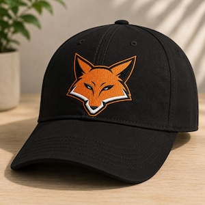 May include: A black baseball cap featuring an embroidered graphic of a fox head on the front panel. The fox design is rendered in shades of orange, white, and black, with sharp, angular lines and a focused expression centered on the crown.