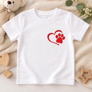 May include: A white short-sleeved t-shirt featuring a red embroidered design on the upper left chest. The design consists of a heart outline that transitions into a paw print. The shirt has a crew neckline and a simple, clean construction.