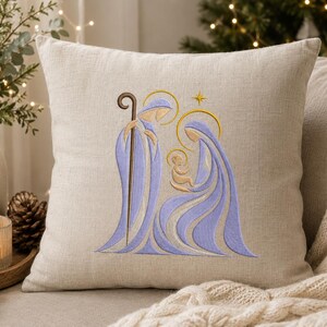 May include: A square beige linen throw pillow featuring an embroidered nativity scene in shades of light purple and gold. The design depicts three figures with golden halos and a small star above, rendered in smooth, flowing lines on the fabric.