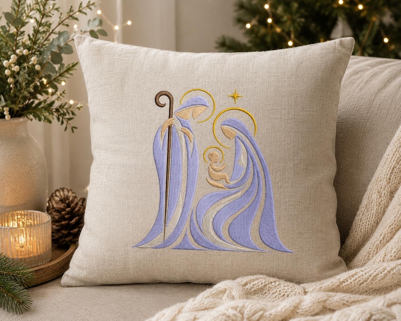 May include: A square beige linen throw pillow featuring an embroidered nativity scene in shades of light purple and gold. The design depicts three figures with golden halos and a small star above, rendered in smooth, flowing lines on the fabric.
