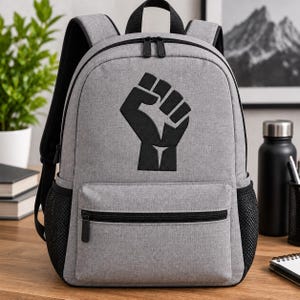 Puede incluir: Una mochila gris claro con un símbolo de puño en alto bordado en negro en el bolsillo delantero. La bolsa incluye cremalleras negras, bolsillos laterales de malla negra y correas ajustables para los hombros. El diseño está centrado.