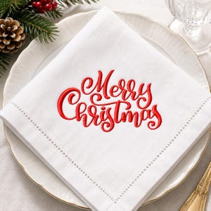 May include: A square white cloth napkin featuring red embroidered cursive text that reads Merry Christmas. The napkin has a decorative hemstitched border and is folded neatly on top of a white ceramic plate with a scalloped edge.