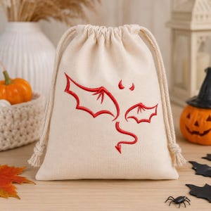 May include: A cream-colored fabric drawstring pouch featuring a red embroidered design of dragon wings, a small tail, and two tiny horns. The bag is made of a plain woven textile and includes two thick, matching cream-colored rope drawstrings.