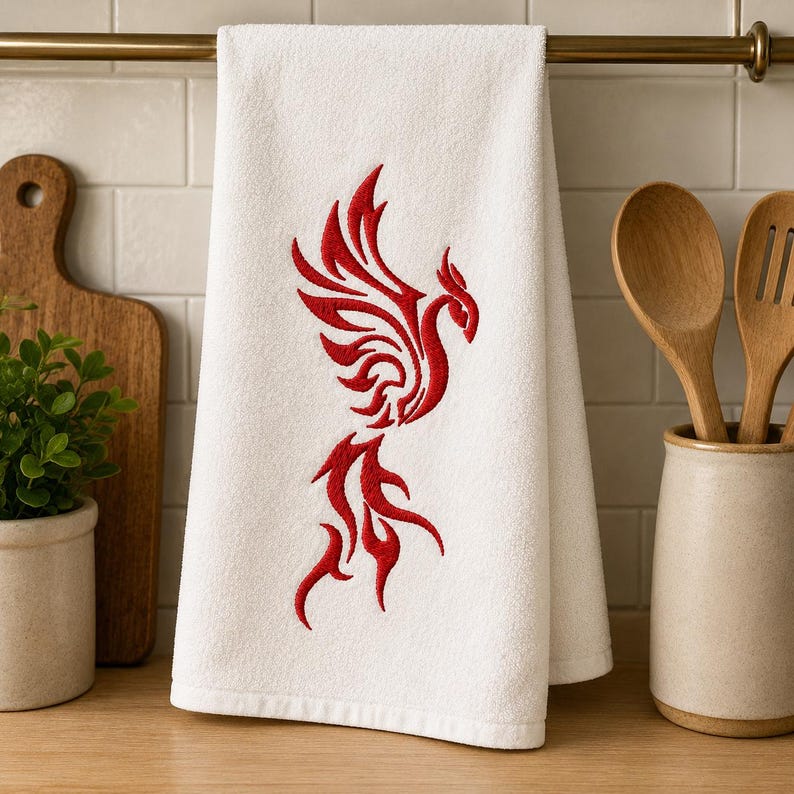 May include: A white kitchen towel featuring a centered red embroidered design of a stylized phoenix with flowing, flame-like feathers. The towel is rectangular with a soft, absorbent texture and hangs vertically from a horizontal metal rod.