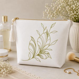 May include: A white zipped pouch featuring an embroidered olive green botanical design of leaves and stems on the front. The pouch includes a white fabric tassel attached to the gold-toned zip pull, providing a clean and simple aesthetic.