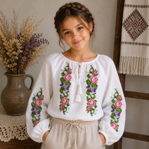 May include: A white long-sleeved blouse featuring vertical floral embroidery in shades of pink, purple, and green. The garment has a round neckline with a central tie closure finished with white tassels and gathered cuffs at the wrists.