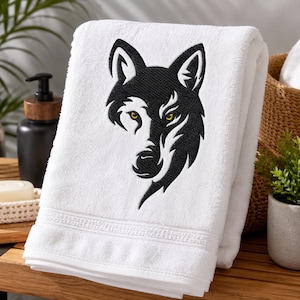 Puede incluir: Una toalla de algodón blanca con un diseño bordado en negro de una cabeza de lobo. El lobo presenta rasgos estilizados y definidos con ojos amarillos penetrantes. El bordado está centrado en la parte frontal de la tela de rizo doblada.