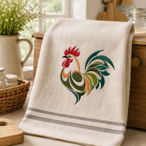 Può includere: Un canovaccio da cucina beige con un gallo ricamato dal piumaggio rosso, arancione, verde e verde acqua. L'asciugamano presenta due strisce orizzontali grigie vicino al bordo inferiore. Il tessuto ha un aspetto intrecciato e testurizzato.