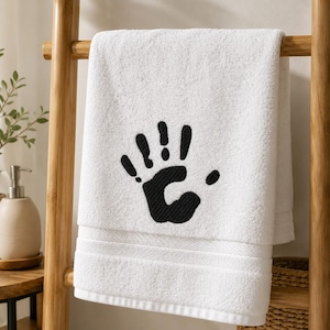 May include: A white cotton bath towel featuring a black embroidered handprint design in the center. The towel has a textured fabric surface with a horizontal ribbed border detail near the bottom edge, showcasing a clean and simple aesthetic.