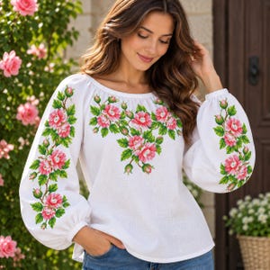 May include: A white long-sleeved blouse featuring an embroidered pattern of pink flowers with green leaves and stems. The embroidery is concentrated around the neckline and extends down the length of both sleeves, which have gathered cuffs.