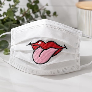 May include: A white pleated face mask featuring a central embroidered graphic of red lips with a light pink tongue sticking out. The design includes a black outline around the lips and tongue, with white elastic ear loops on both sides.
