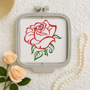 May include: A square gray embroidery hoop holding a white fabric panel featuring a machine-embroidered design of a single red rose with green leaves and stems. The rose is outlined in bold red thread with green accents on the foliage below.