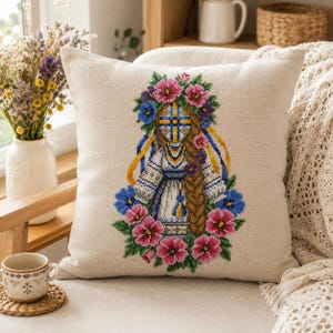 May include: A square beige throw pillow featuring a cross-stitch embroidery of a figure with a long braided hairstyle, wearing a white garment with blue and yellow patterns, surrounded by a vibrant wreath of pink, purple, and blue flowers.