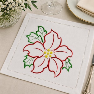 May include: A square white fabric napkin featuring an embroidered poinsettia design in the center. The flower has red outlines, yellow centers, and green holly leaves. The napkin has a decorative hemstitched border along all four edges.