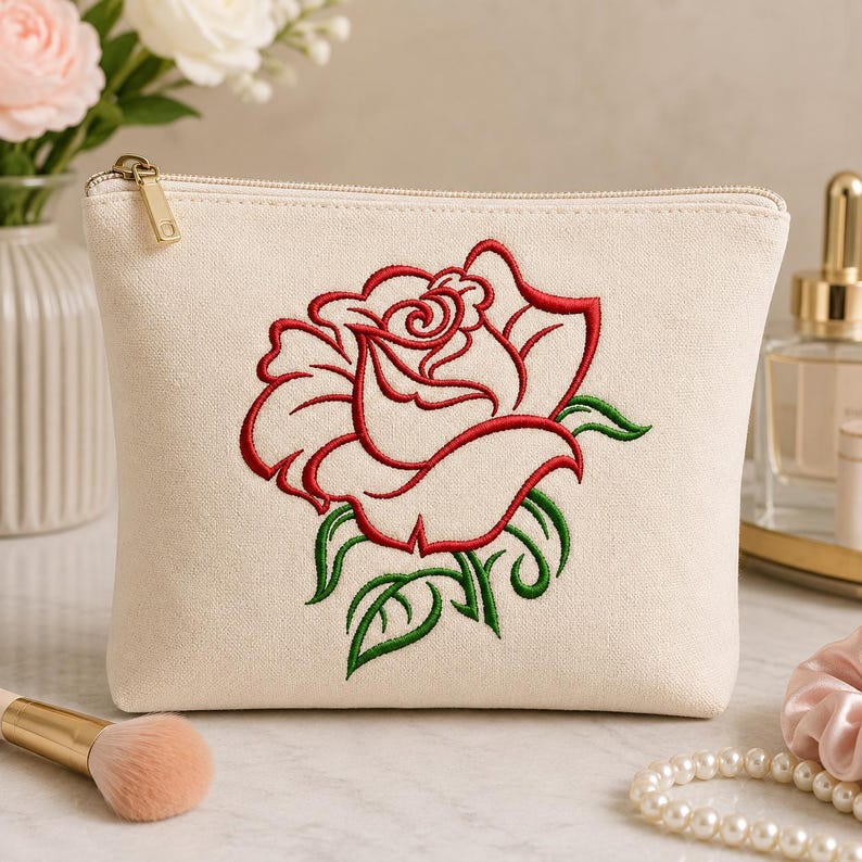 May include: A cream-colored canvas zippered pouch featuring a large, centered embroidery of a red rose with green leaves and stems. The pouch has a gold-toned metal zipper pull and a rectangular shape with rounded top corners.