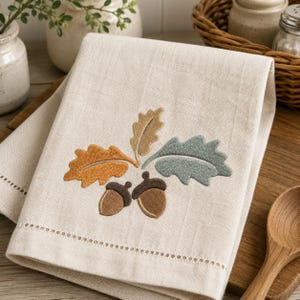 May include: A folded off-white linen kitchen towel featuring an embroidered design of two brown acorns and three oak leaves in shades of golden yellow, tan, and muted sage green. The towel has a decorative hemstitch detail along the bottom edge.