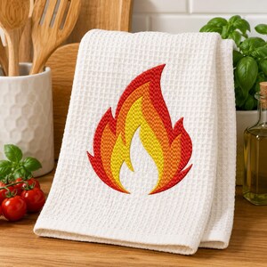 May include: A white waffle-weave kitchen towel featuring a centered embroidered flame design. The flame is composed of layered sections in vibrant shades of red, orange, and yellow, creating a stylized fire graphic on the textured fabric surface.