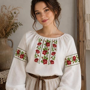 Könnte beinhalten: Eine weiße Langarmbluse mit Rundhalsausschnitt und aufwendiger Kreuzstichstickerei. Das Design zeigt leuchtend rote Rosen mit grünen Blättern und Stielen, die vertikal auf der Brust und horizontal an den Ärmeln angeordnet sind.