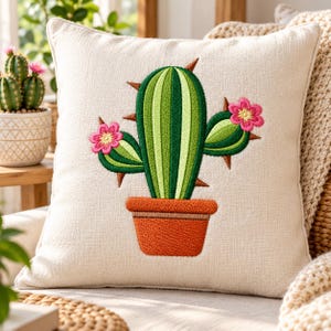 Può includere: Un cuscino quadrato color bianco sporco con un ricamo centrale raffigurante un cactus verde in un vaso arancione. Il cactus presenta strisce verticali in varie tonalità di verde, spine marroni e due fiori rosa brillante sui bracci laterali.