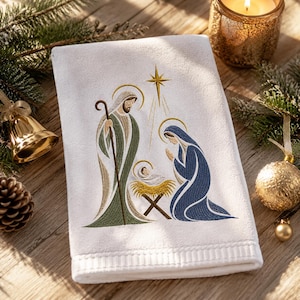 May include: A white hand towel featuring an embroidered nativity scene. The design depicts a figure in a green robe holding a staff, a figure in a blue robe kneeling, and a baby in a manger, all beneath a gold star with radiating light rays.