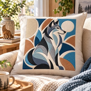 Puede incluir: Un cojín cuadrado con un diseño geométrico de lobo bordado en tonos azules, negros y bronceados sobre una tela de color crema. El lobo está representado de perfil sobre un fondo de formas circulares y curvas.