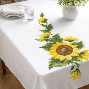 May include: White tablecloth with a cross-stitched sunflower design in yellow, green, and brown. The floral pattern is on the corner of the tablecloth. A glass of water, plates, and a vase of flowers are on the table.