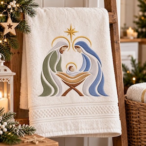 Puede incluir: Una toalla de baño de color crema con una escena de natividad bordada. El diseño muestra dos figuras con túnicas verdes y azules sobre un pesebre central con un bebé, bajo una estrella dorada. La toalla tiene un borde con textura.