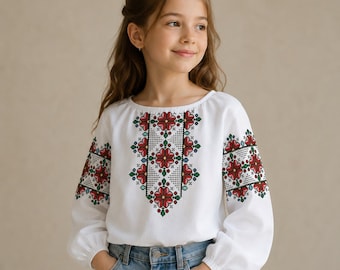 May include: A white long-sleeved blouse featuring a central vertical panel and sleeve accents with a red, green, and black geometric cross-stitch floral pattern. The garment has a rounded neckline and gathered cuffs, paired with light blue denim jeans.