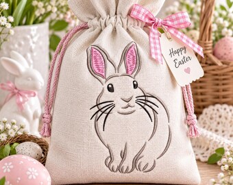 Può includere: Un sacchetto regalo in lino beige con coulisse, caratterizzato da un coniglio ricamato con orecchie dai dettagli rosa. Il sacchetto è chiuso da cordoncini intrecciati rosa e decorato con un nastro a quadretti rosa e bianco su un'etichetta.