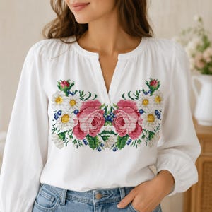 May include: A white long-sleeved blouse featuring a split neckline and a colorful cross-stitch embroidery pattern on the chest. The design displays two large pink roses surrounded by white daisies, small blue flowers, and green leaves.
