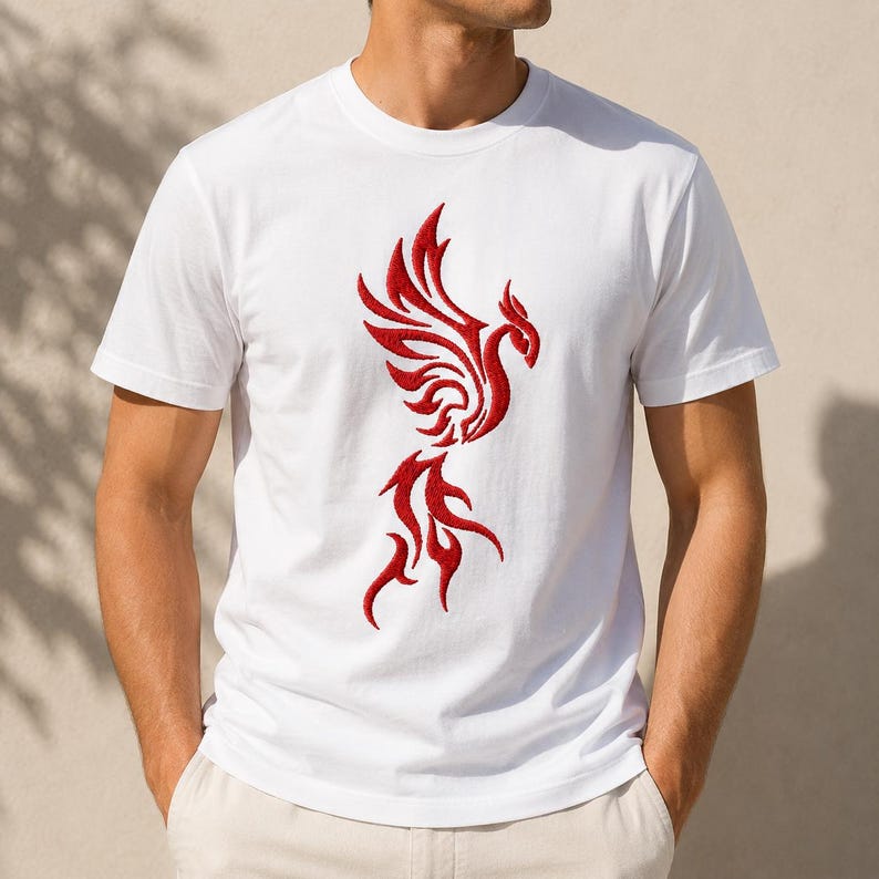 May include: A white short-sleeved t-shirt featuring a prominent red embroidered phoenix design centered on the chest. The phoenix is depicted with stylized, flowing wings and a long, flame-like tail, creating a bold contrast against the plain white fabric.