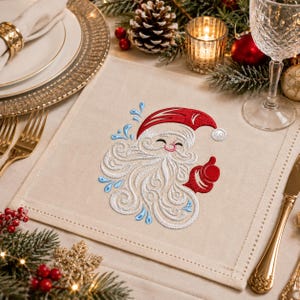 May include: A square beige fabric napkin featuring an embroidered design of a smiling Santa Claus face with a swirling white beard, a red hat, and light blue decorative accents. The embroidery is centered on the napkin with a stitched border.