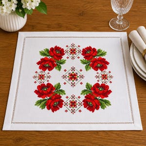 May include: A square white fabric placemat featuring a central cross-stitch pattern of four clusters of vibrant red poppies with green leaves, arranged in a circular formation with small geometric red and green floral motifs in between.