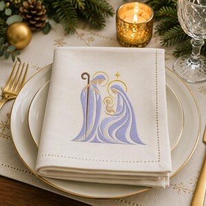 May include: A folded off-white linen napkin featuring an embroidered nativity scene in shades of light purple and gold. The design depicts three figures with halos and a staff, centered on the fabric which has a decorative stitched border.