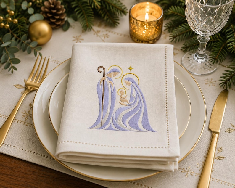 May include: A folded off-white linen napkin featuring an embroidered nativity scene in shades of light purple and gold. The design depicts three figures with halos and a staff, centered on the fabric which has a decorative stitched border.