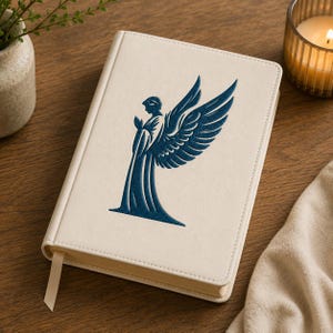 May include: A cream-colored hardcover journal featuring a dark blue embossed graphic of an angel with large wings on the front cover. The notebook includes a matching cream-colored ribbon bookmark extending from the bottom edge of the spine.