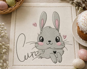 Può includere: Un tovagliolo di stoffa quadrato color bianco sporco con il ricamo di un coniglio grigio con guance e orecchie rosa. La parola "Cute" è ricamata in corsivo nero a sinistra del coniglio, accompagnata da piccoli cuori rosa e neri.